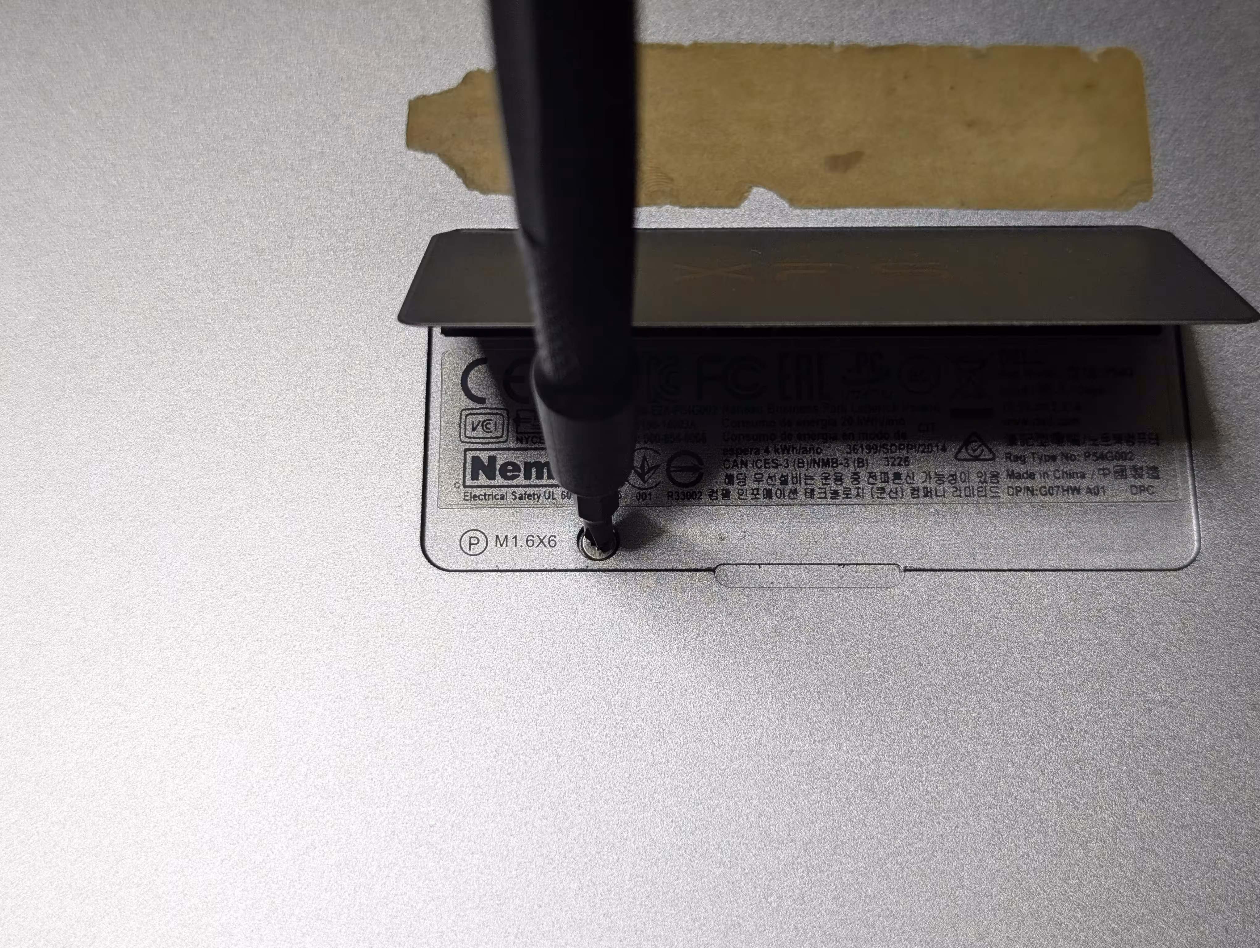 A flap in the middle of the laptop's bottom cover is partially opened, with a screwdriver keeping it from closing. The screwdriver is placed on top of a screw underneath the flap.