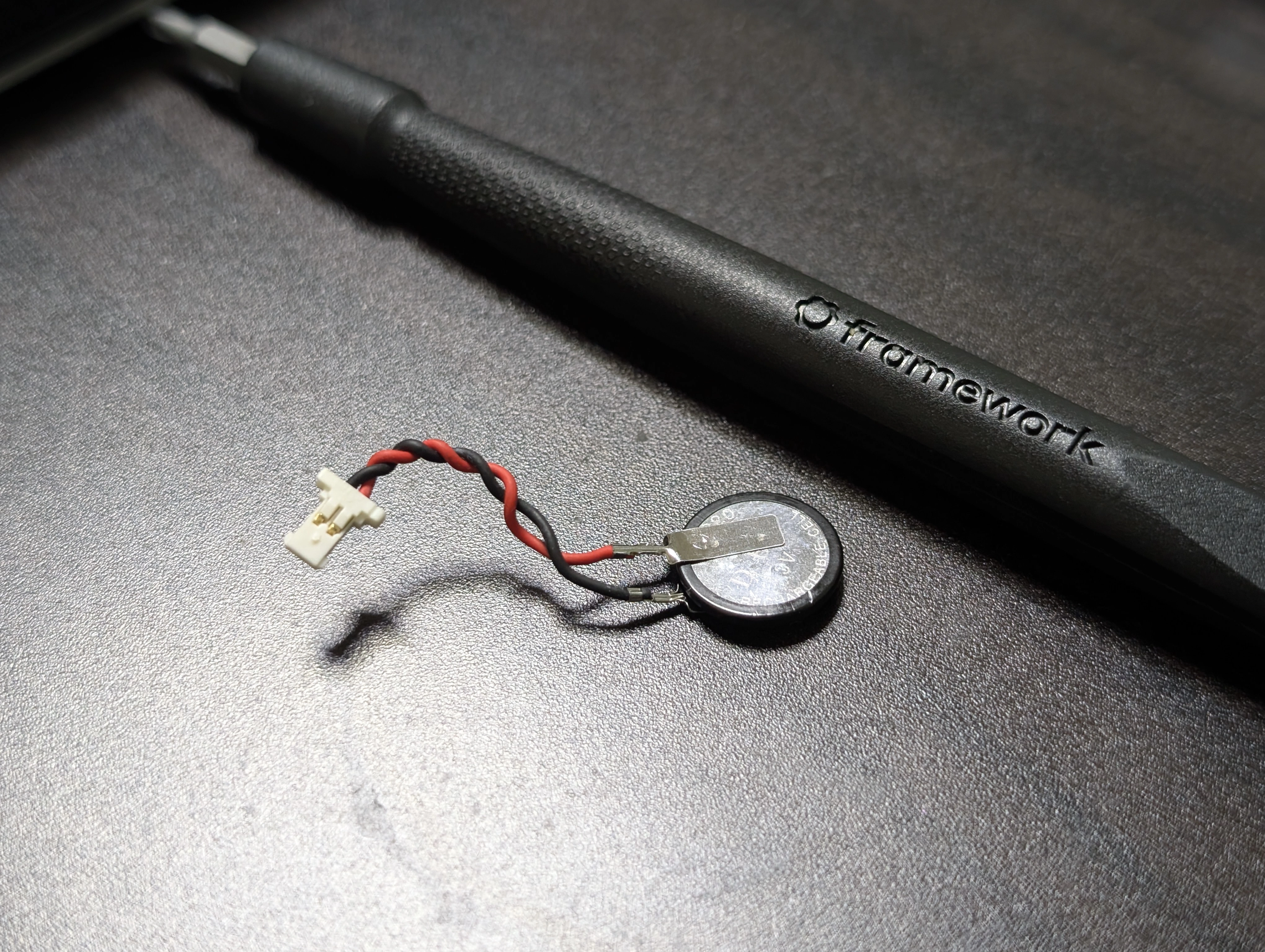 The disconnected RTC battery, with its black plastic wrap peeled off, next to a Framework Screwdriver